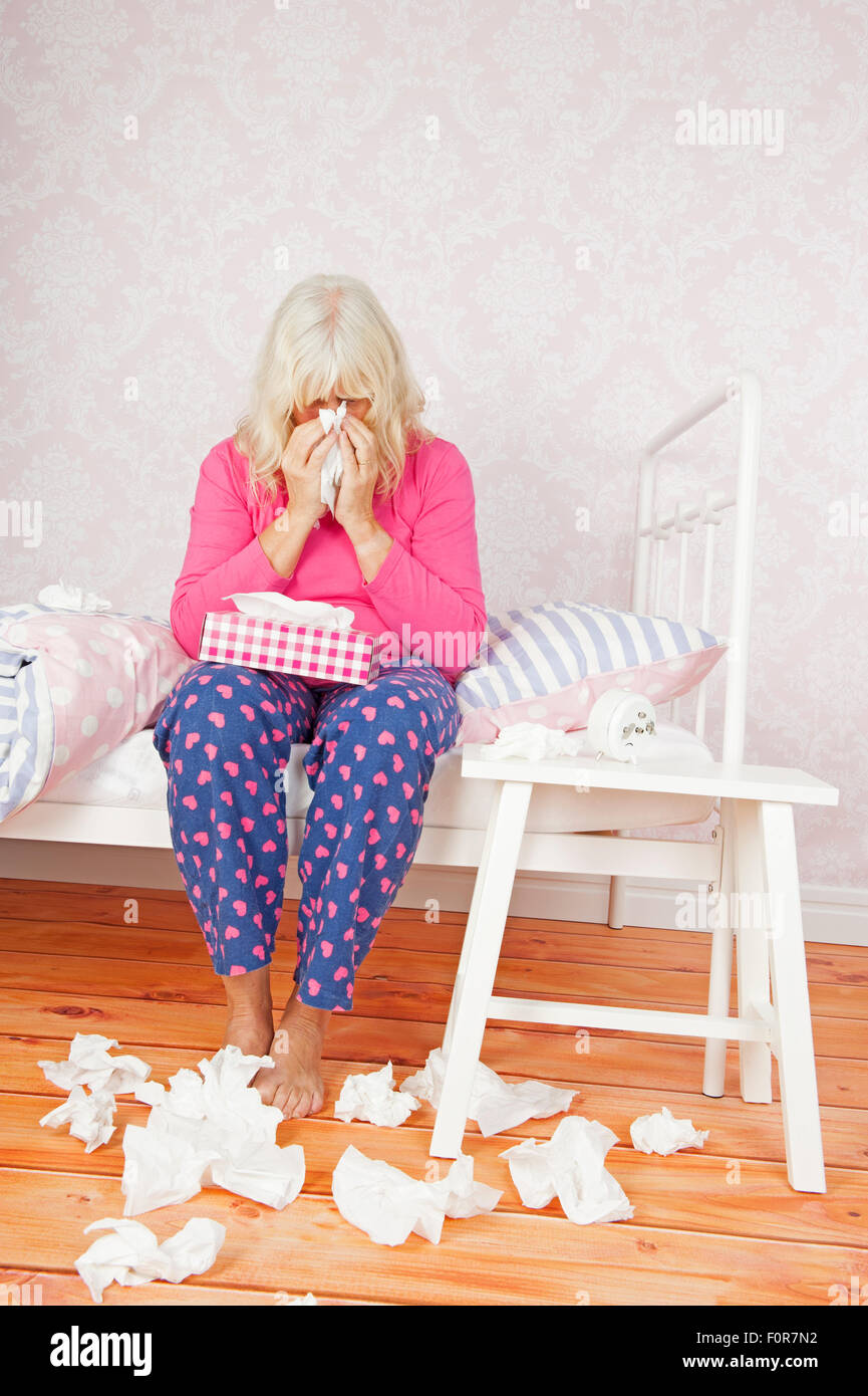 Traurige Frau mit rosa Pyjama und Geweben, die ihre Nase weht, sitzend auf Bett Stockfoto