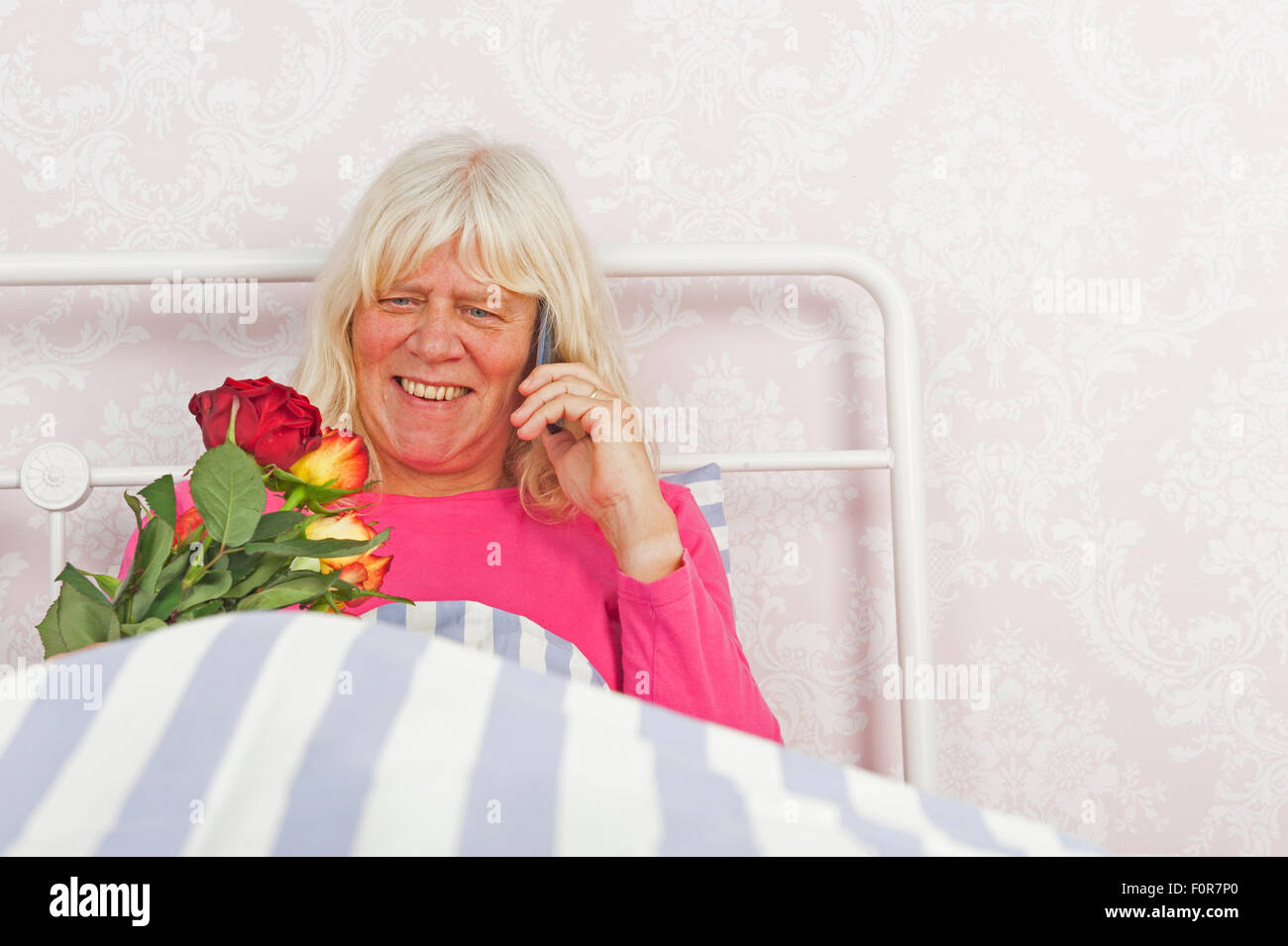 Glückliche Frau in rosa Pyjama im Bett mit einem Strauß Rosen sitzen und reden am Telefon Stockfoto