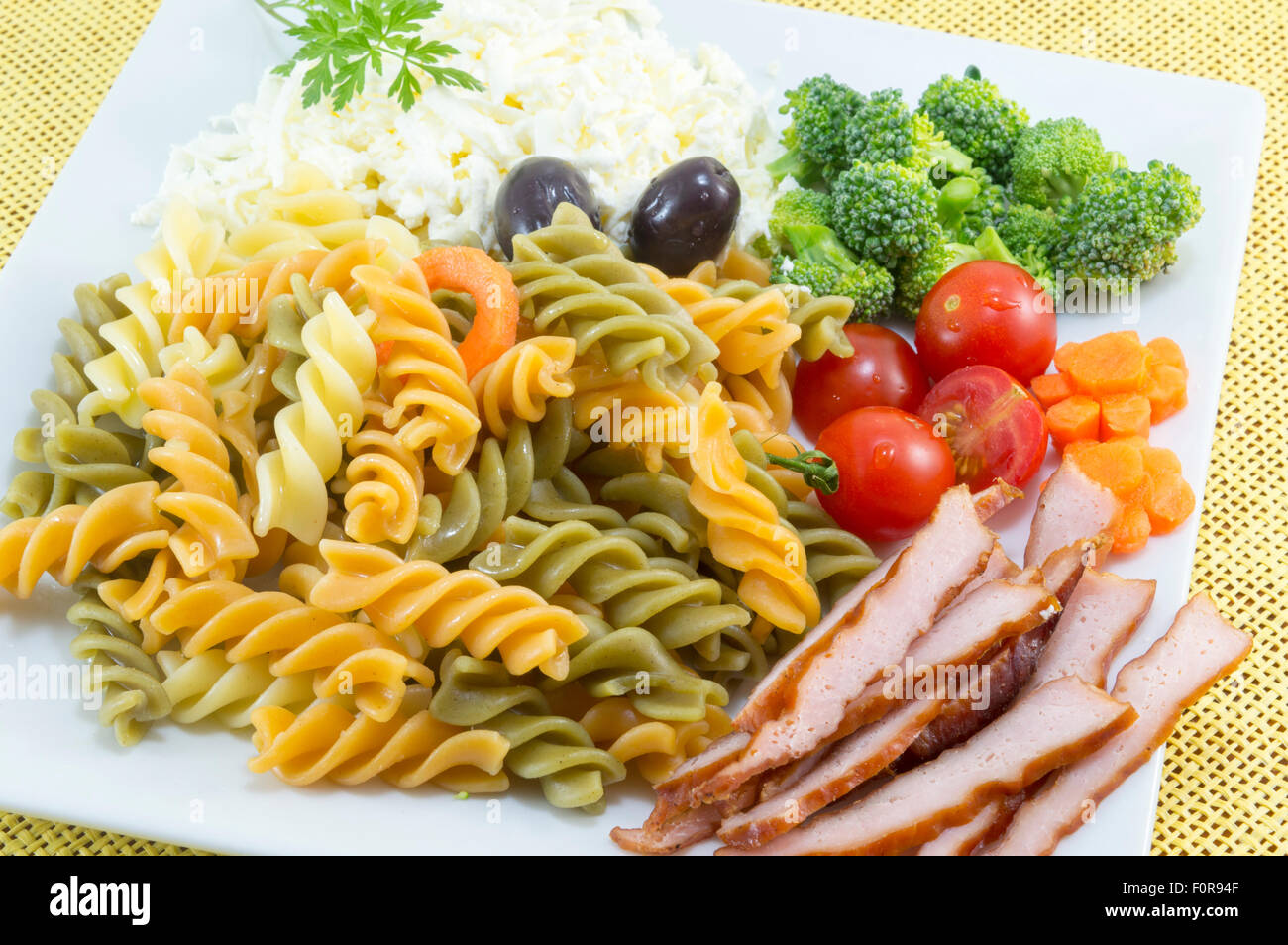 Farbige Pasta serviert auf einem weißen Teller mit Käse, Oliven, Brokkoli, Cherry-Tomaten und geräuchertem Fleischscheiben Stockfoto