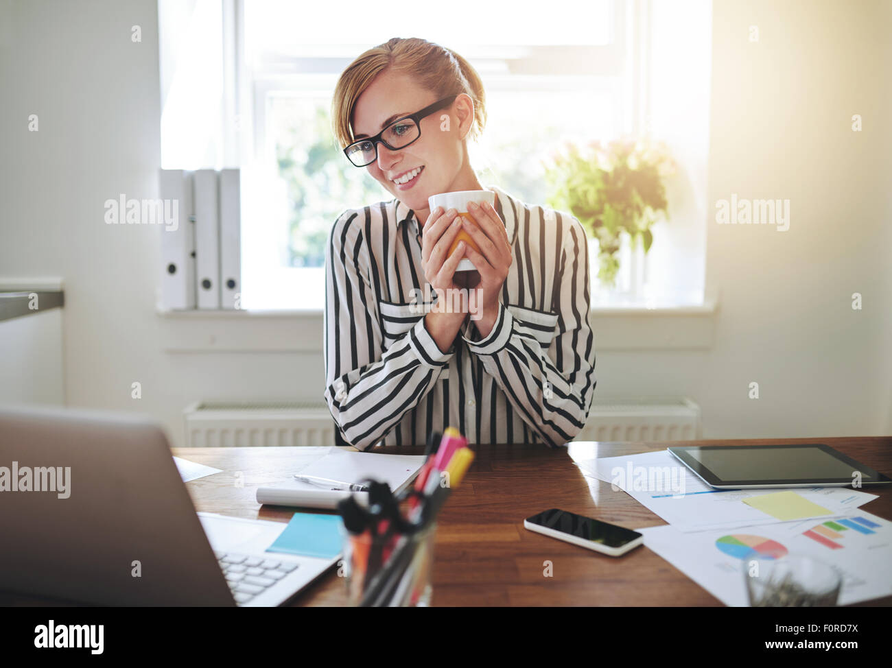 Glücklich erfolgreiche Geschäftsfrau eine Pause, eine Tasse Kaffee zu trinken, wie sie Informationen auf ihrem Laptop-Bildschirm mit einer Smil liest Stockfoto
