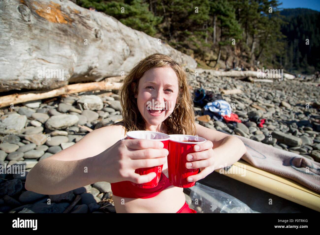 Junge Frau sitzt am felsigen Strand, hält Getränke, lächelnd, Short Sands Beach, Oregon, USA Stockfoto Junge Frau sitzt am felsigen Strand, hält Getränke, lächelnd, Short Sands Beach, Oregon, USA Stockfoto