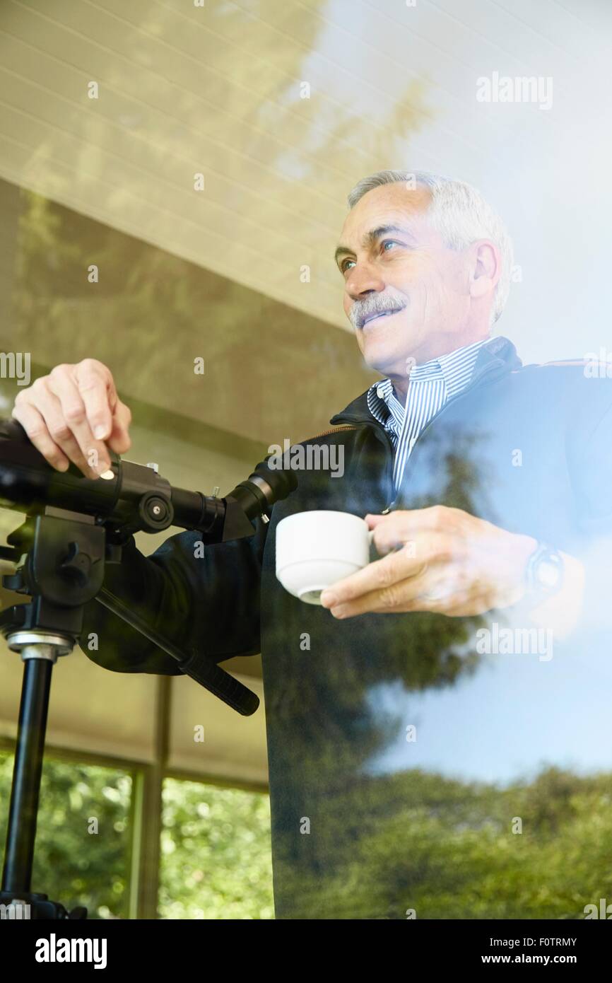 Ältere Mann zu Hause Kaffee trinken, mit Teleskop durch Fenster Stockfoto