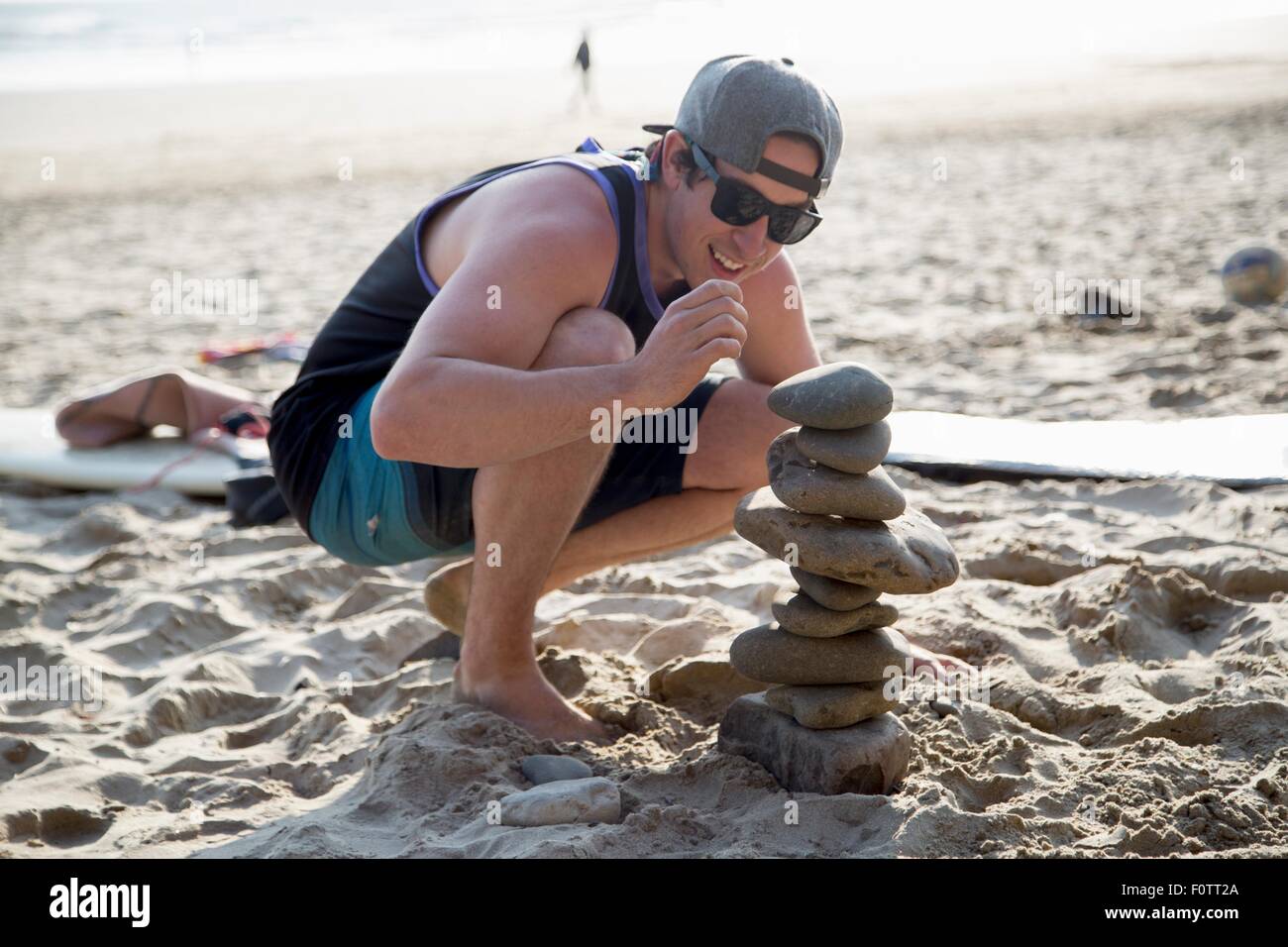 Junger Mann kauert neben einem gestapelten Steinen am Strand Stockfoto