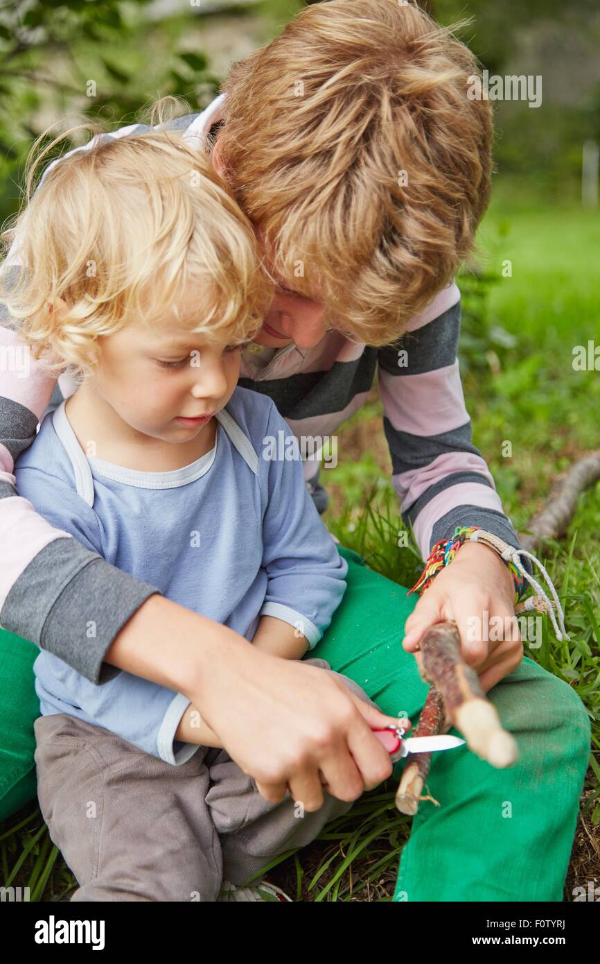 Junge sitzt mit jüngeren Bruder mit einem Taschenmesser auf Zweig im Garten Stockfoto