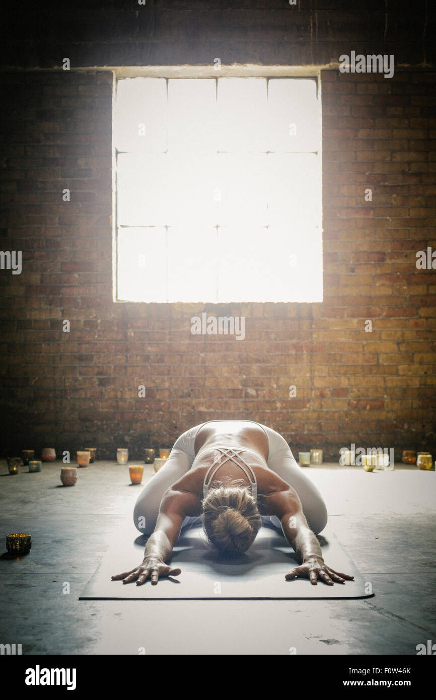 Eine blonde Frau in einem weißen crop Top und Leggings, bücken auf einer Yogamatte, umgeben von Kerzen, Yoga zu tun. Stockfoto