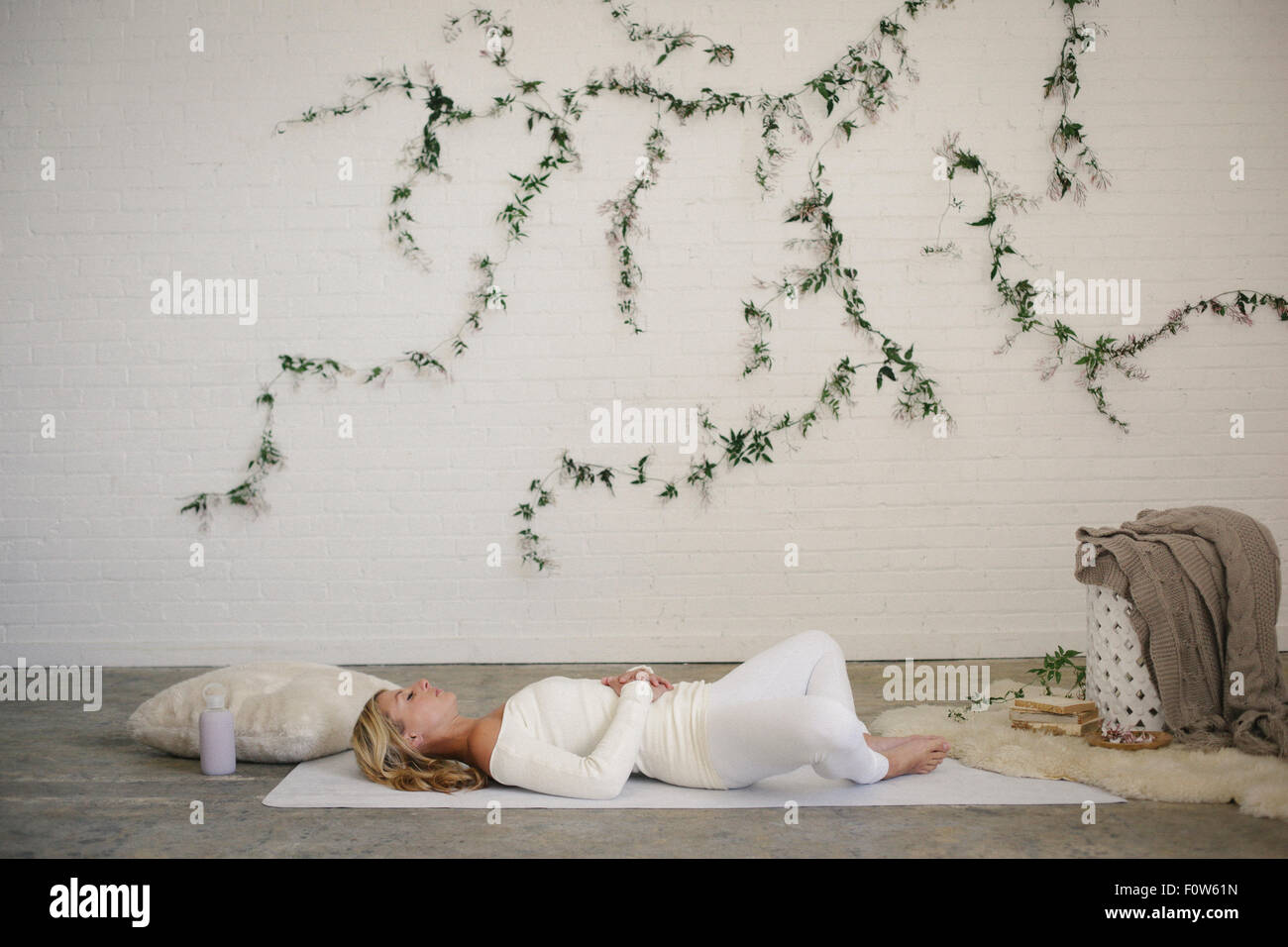 Eine blonde Frau in einem weißen Trikot und Leggings, liegend auf einer weißen Matte in einem Raum. Eine Schlingpflanze Pflanze an der Wand hinter ihr. Stockfoto