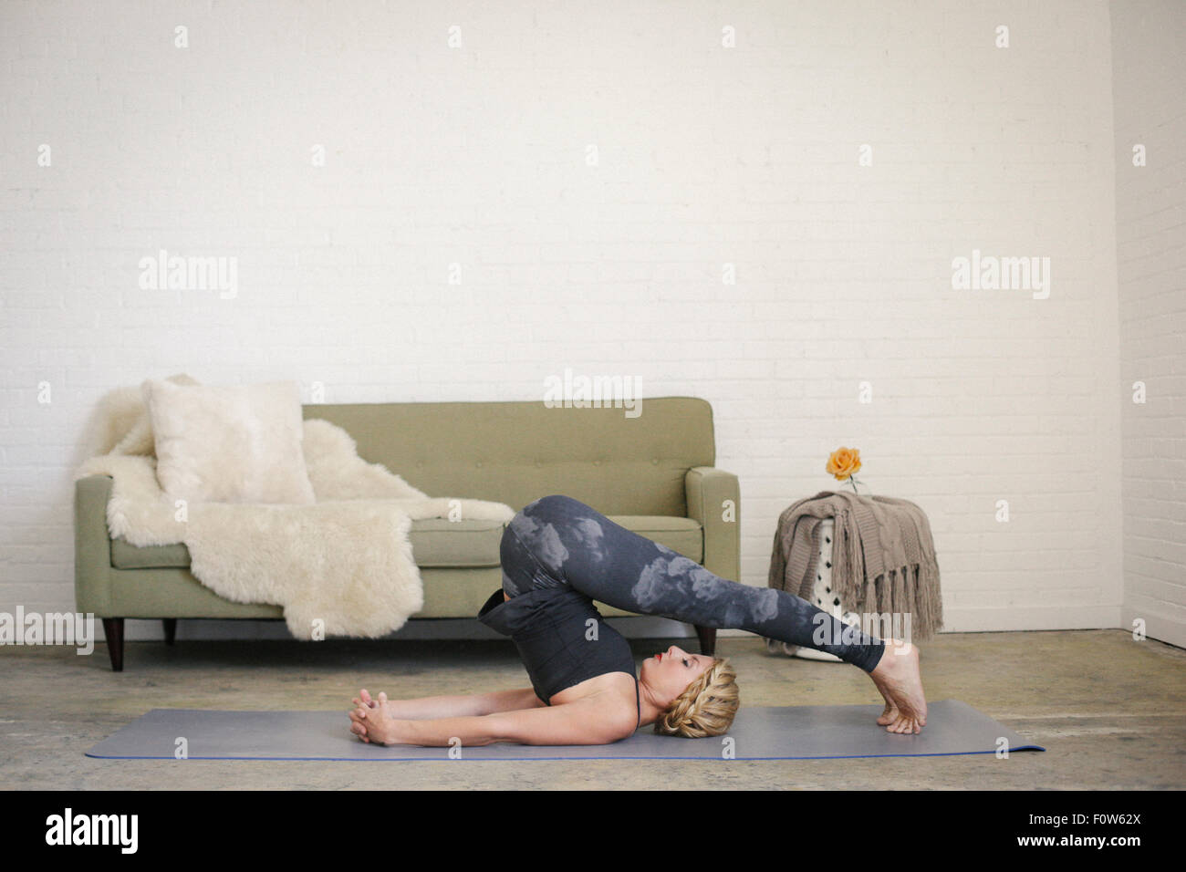Eine blonde Frau in einem schwarzen Anzug und Leggings, liegend auf einer Yogamatte in einem Raum, Yoga, ihre Beine, beugte sich über den Kopf zu tun. Stockfoto