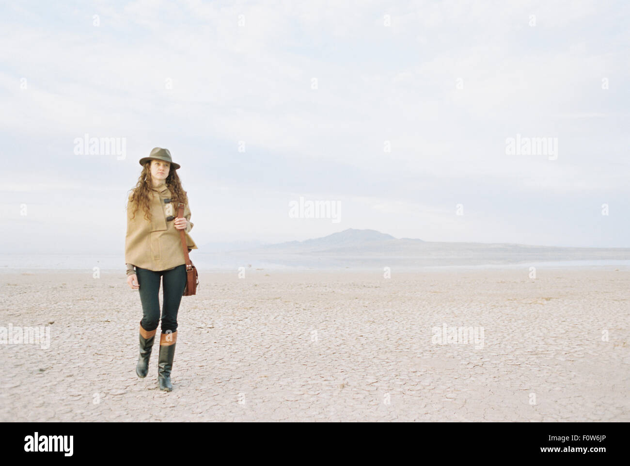 Eine Frau mit braunen, langen und lockigen Haar zu Fuß durch eine Wüstenebene, einen Hut und eine Leder-Tragetasche. Stockfoto