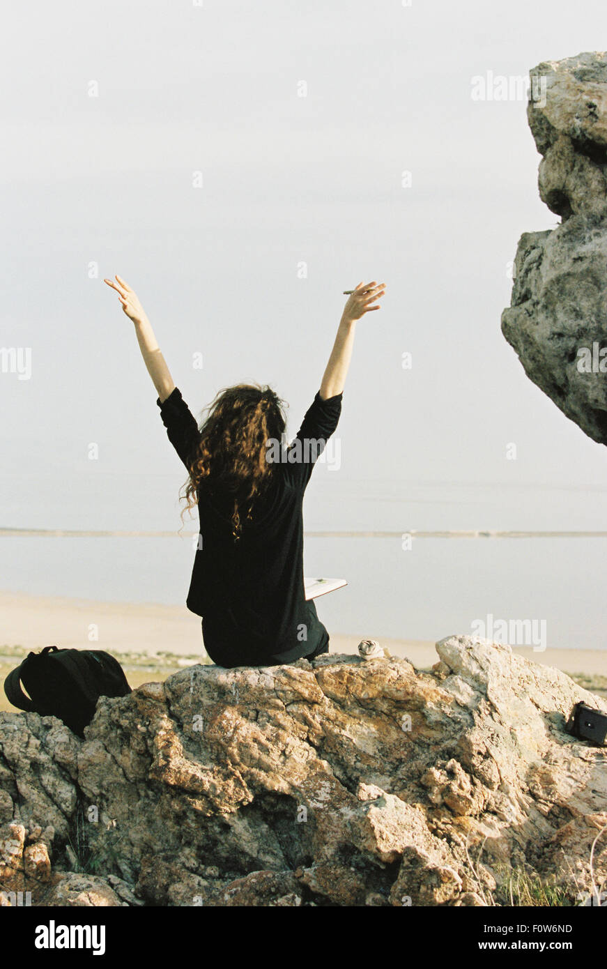 Eine Frau sitzt auf einem Felsen mit ihren Armen angehoben, Blick auf Wasser Stockfoto