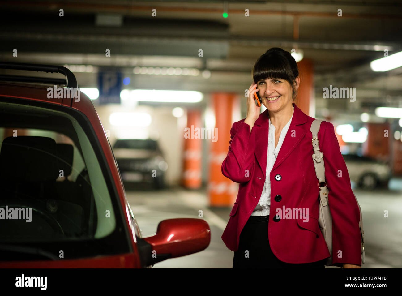 Reife Geschäftsfrau tragen rote Jacke aufrufenden Handy im stehen bei ihrem Auto lächelnd Stockfoto