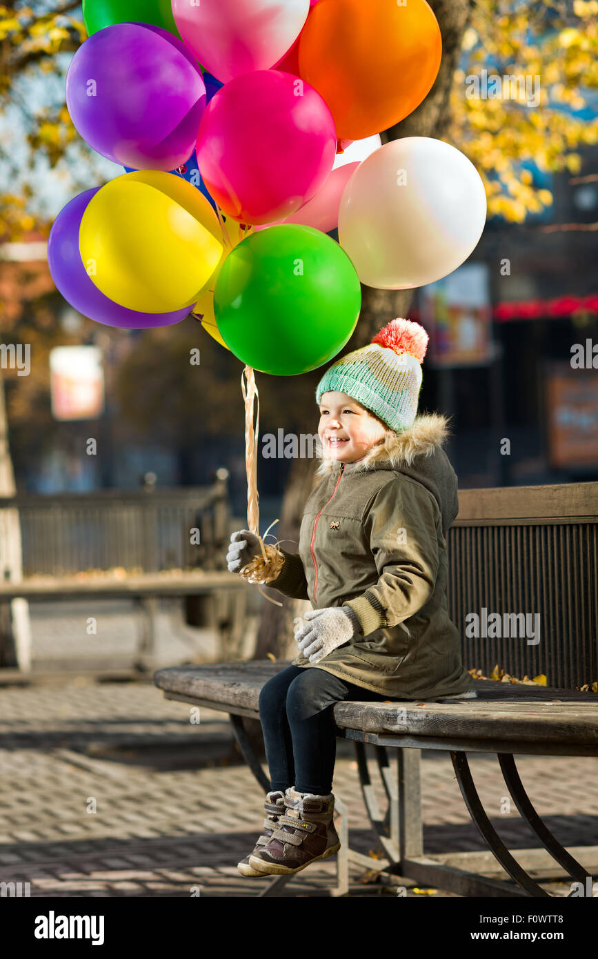 glückliches kleines Kind sitzen auf Bank mit Luftballons und lachen Stockfoto