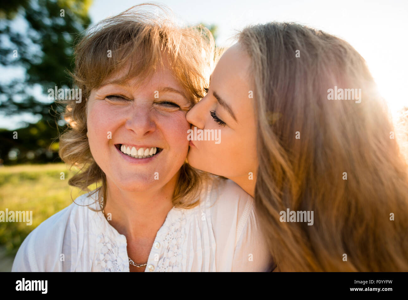 Teenager-Tochter küssen ihre Mutter im Freien in der Natur mit Sonne im Hintergrund, Weitwinkel Stockfoto
