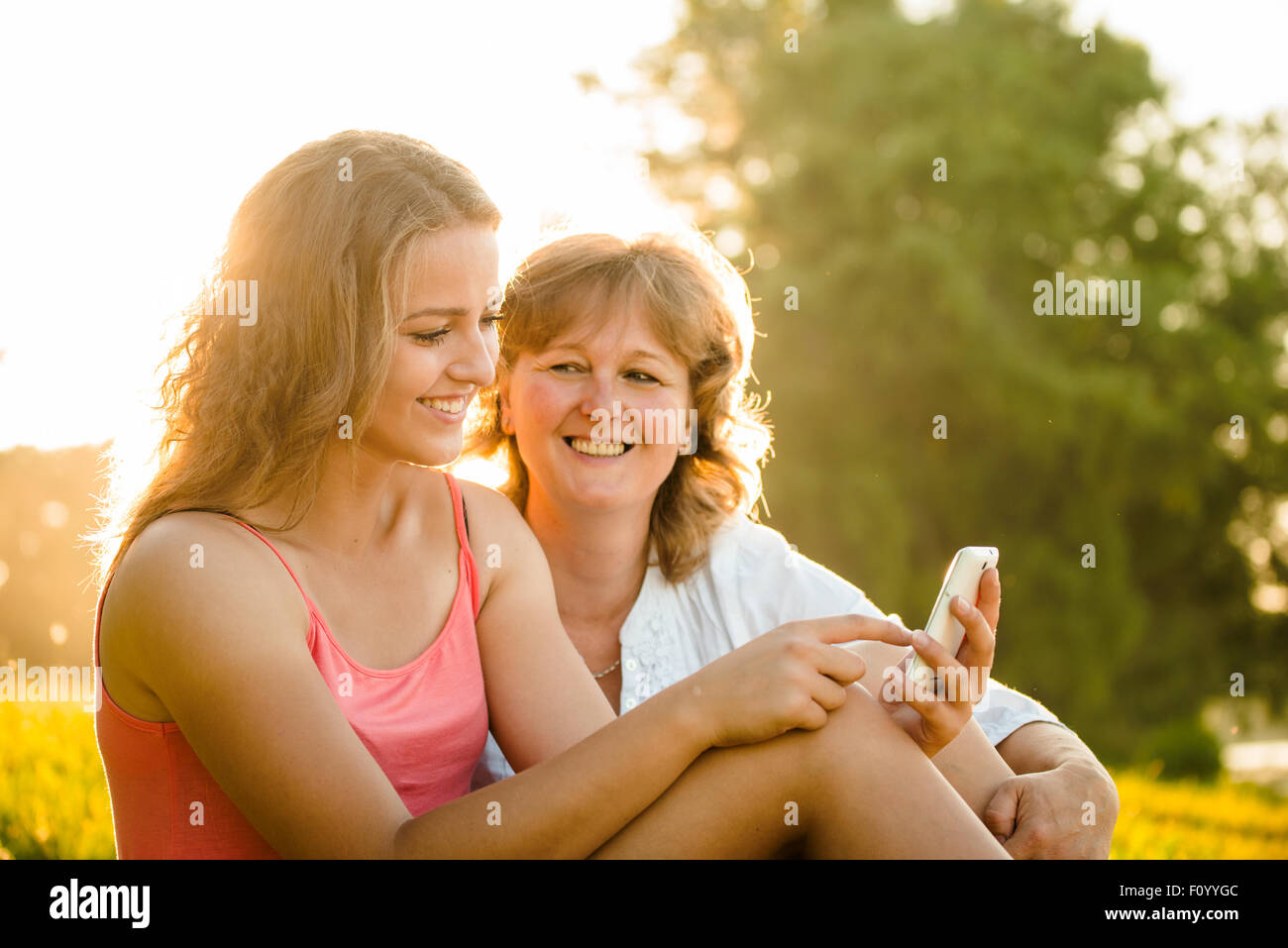 Junges Mädchen zeigt ihre Mutter Fotos auf Handy im Freien in der Natur mit Sonnenuntergang im Hintergrund Stockfoto