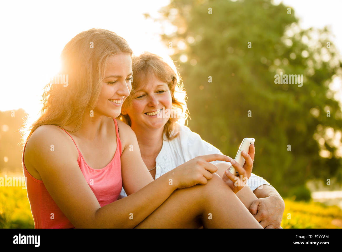 Junges Mädchen zeigt ihre Mutter Fotos auf Handy im Freien in der Natur mit Sonnenuntergang im Hintergrund Stockfoto
