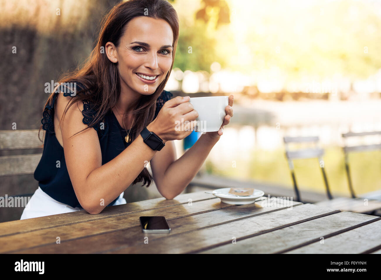 Porträt von schöne junge Frau sitzt an einem Tisch mit einer Tasse Kaffee in der hand beim Café Kamera Lächeln betrachten. Stockfoto