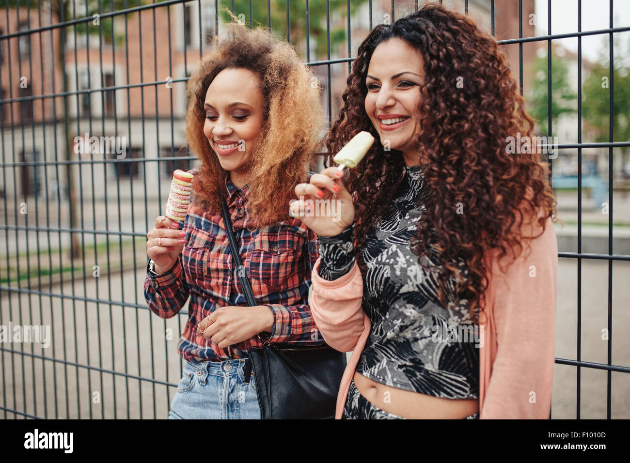 Fröhliche junge Mädchen essen Süßigkeiten Eis. Zwei junge Frauen stehen gegen einen Zaun lächelnd, im Freien. Stockfoto