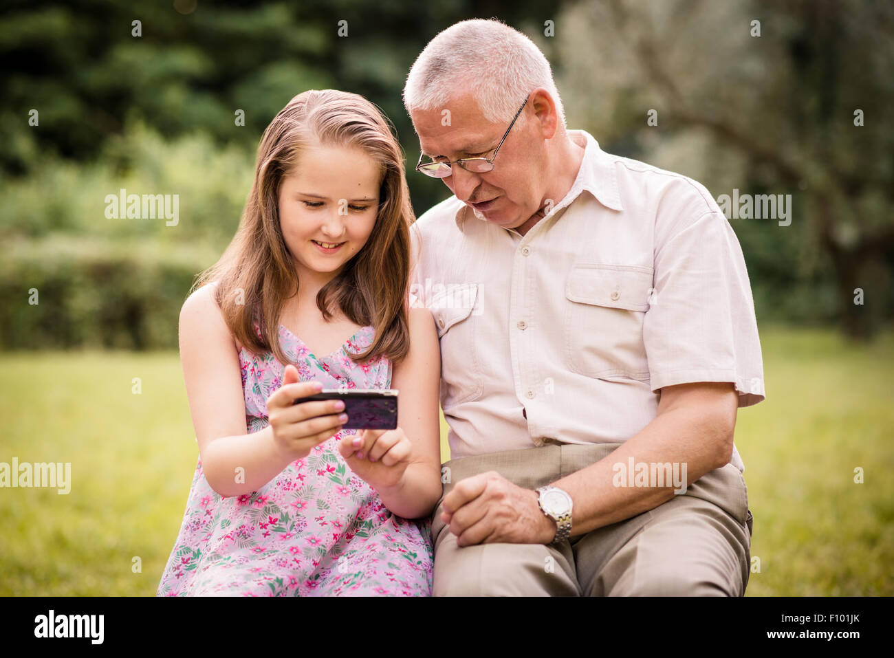 Ältere Mann mit seinem Enkel suchen gemeinsam auf Fotos in Smartphone - draußen in der Natur Stockfoto