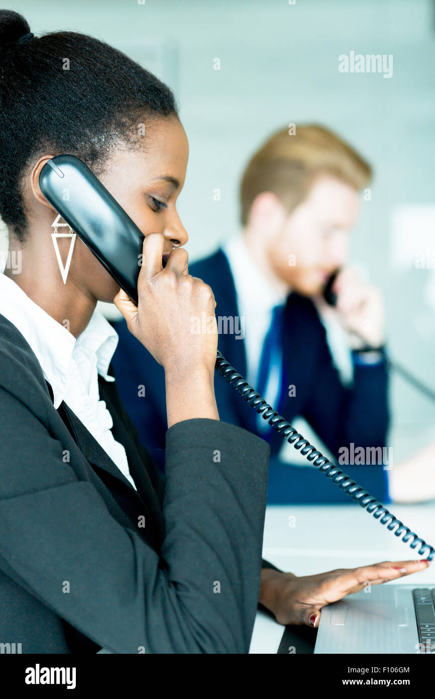 Eine schöne, schwarze, junge Frau arbeitet in einem Callcenter in einem Büro mit ihrem roten Haird Partner am anderen Ende des Schreibtisches ta Stockfoto