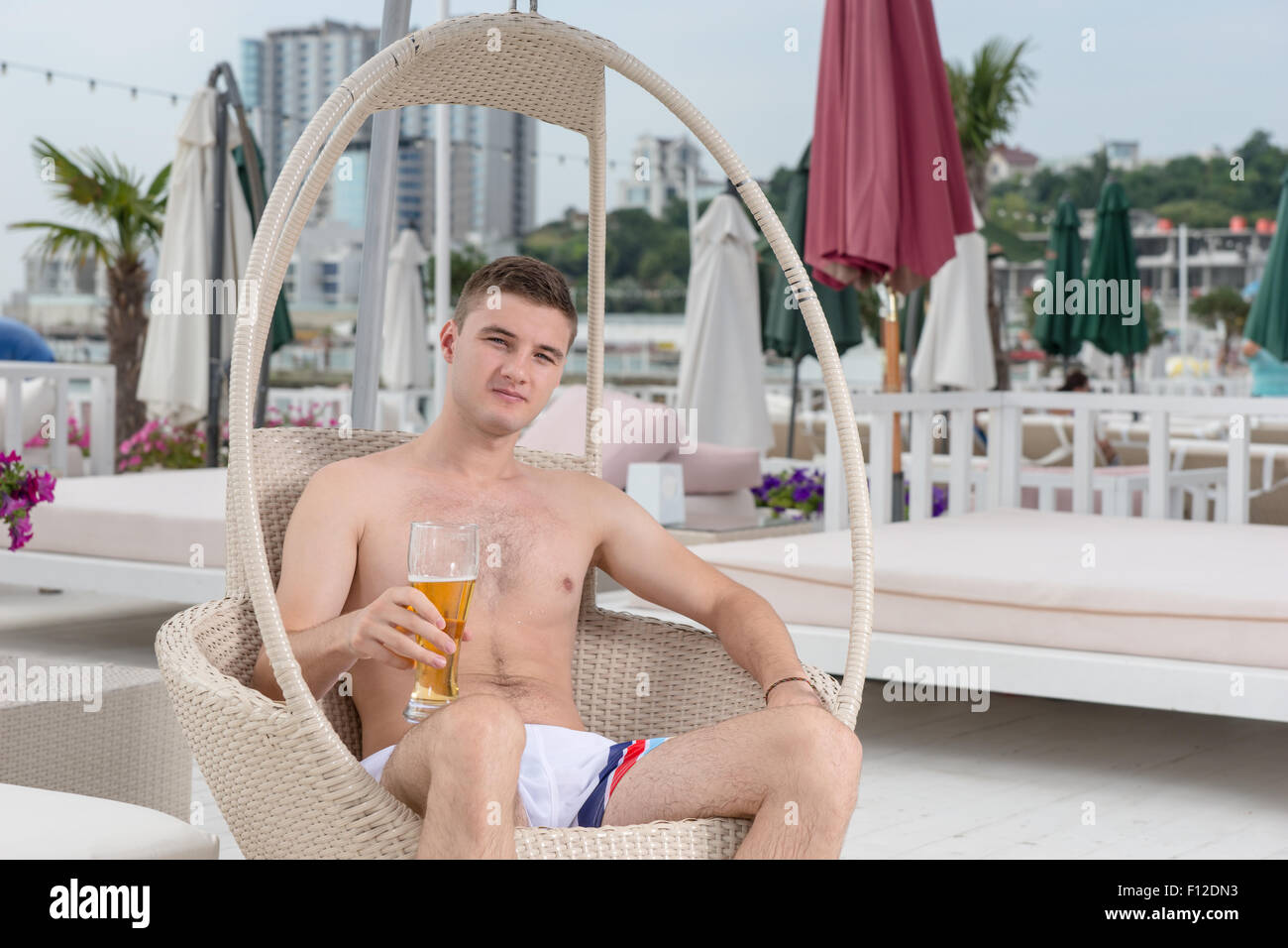 Porträt des jungen Mann im Badeanzug in Korbstuhl mit hohen Glas kaltes Bier auf dem Deck des Luxus-Ferien-Resort direkt am Meer entspannen. Stockfoto