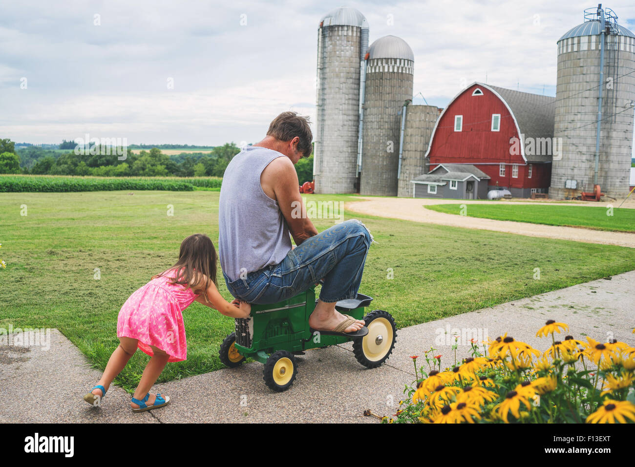 Mädchen schiebt ihren Vater auf einem Spielzeugtraktor auf einer Farm, USA Stockfoto