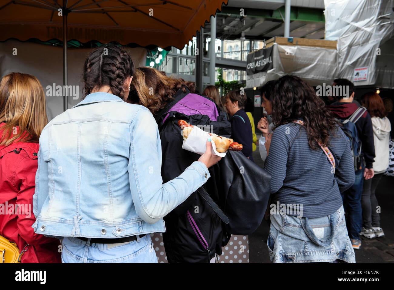 Junge Frau in einer Masse von Käufern holding Essen hotdog von bun Borough Market Southwark, South London UK KATHY DEWITT Stockfoto