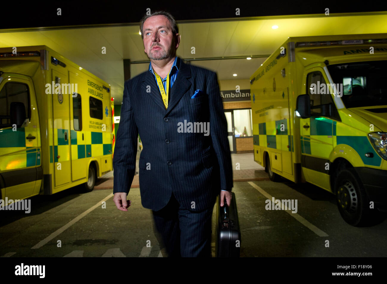 Eine männliche Out-of-Stunden Bereitschaftsarzt, hält Medizintasche und Handy, vorbei an einer Reihe von Krankenwagen in einem Krankenhaus Großbritannien GP Stockfoto