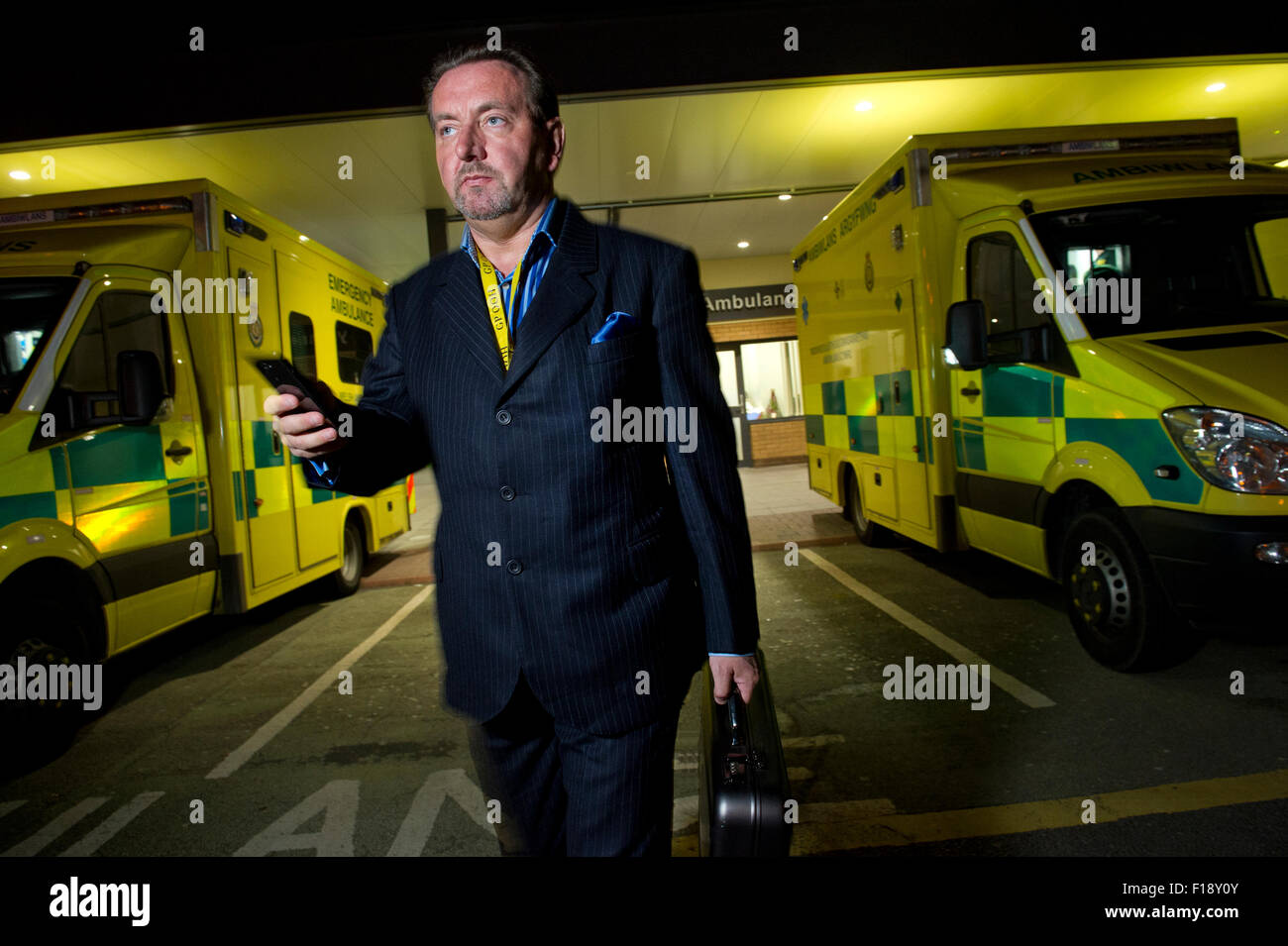 Eine männliche Out-of-Stunden Bereitschaftsarzt, hält Medizintasche und Handy, vorbei an einer Reihe von Krankenwagen in einem Krankenhaus Großbritannien GP Stockfoto