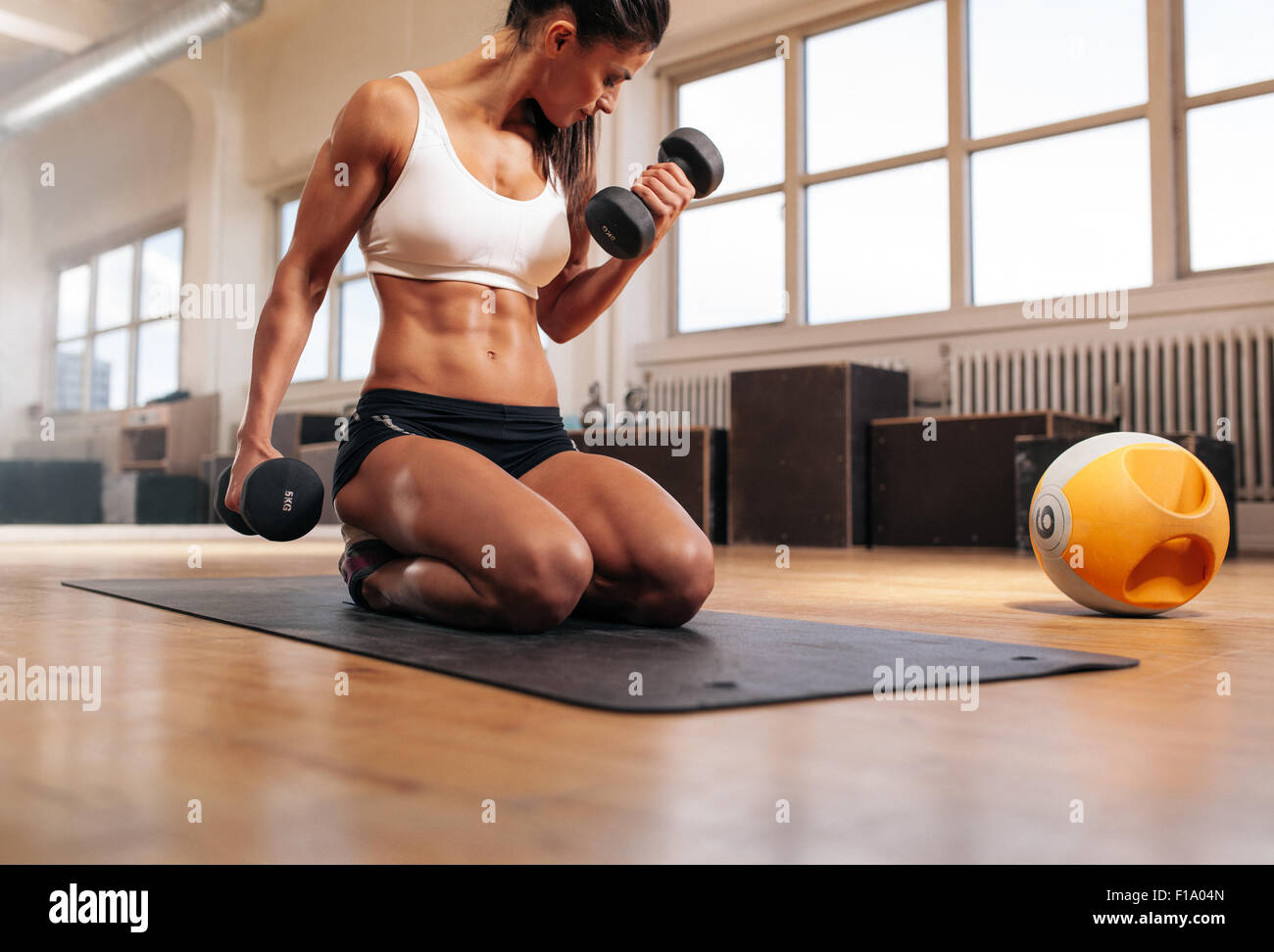 Körperlich fit Frau im Fitnessstudio Hanteln zur Stärkung der Arme und Bizeps heben. Muskulöse Frau sitzen auf Übung Matte Optik Stockfoto