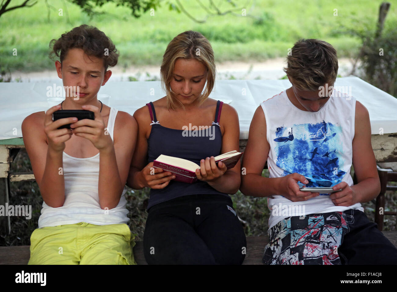 Lago di Bolsena, Italien, spielen die Jungs mit ihren Smartphones während eines Mädchens in einem Buch lesen Stockfoto