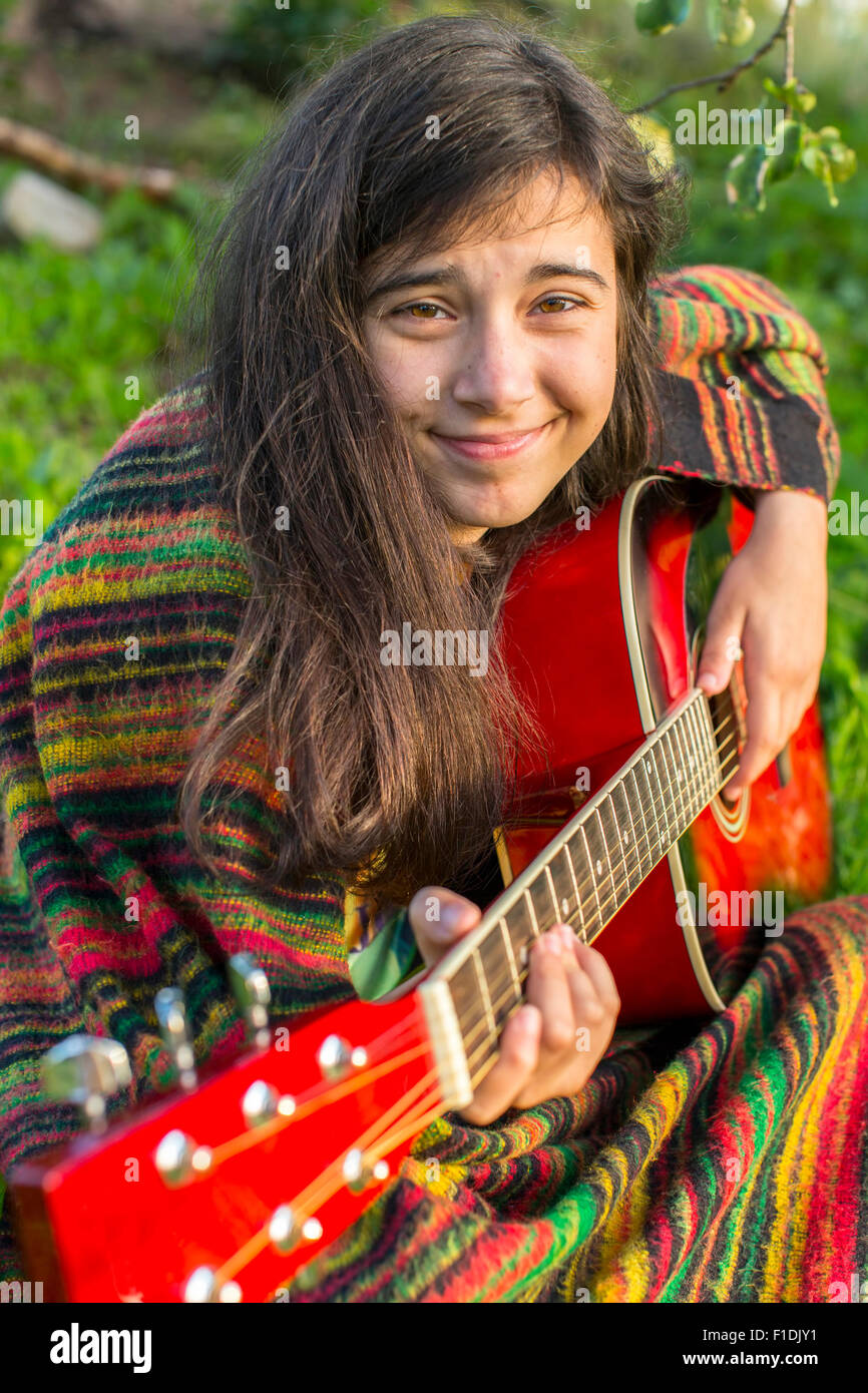 Teenager-Mädchen in Poncho spielt Gitarre beim Sitzen im Freien. Stockfoto