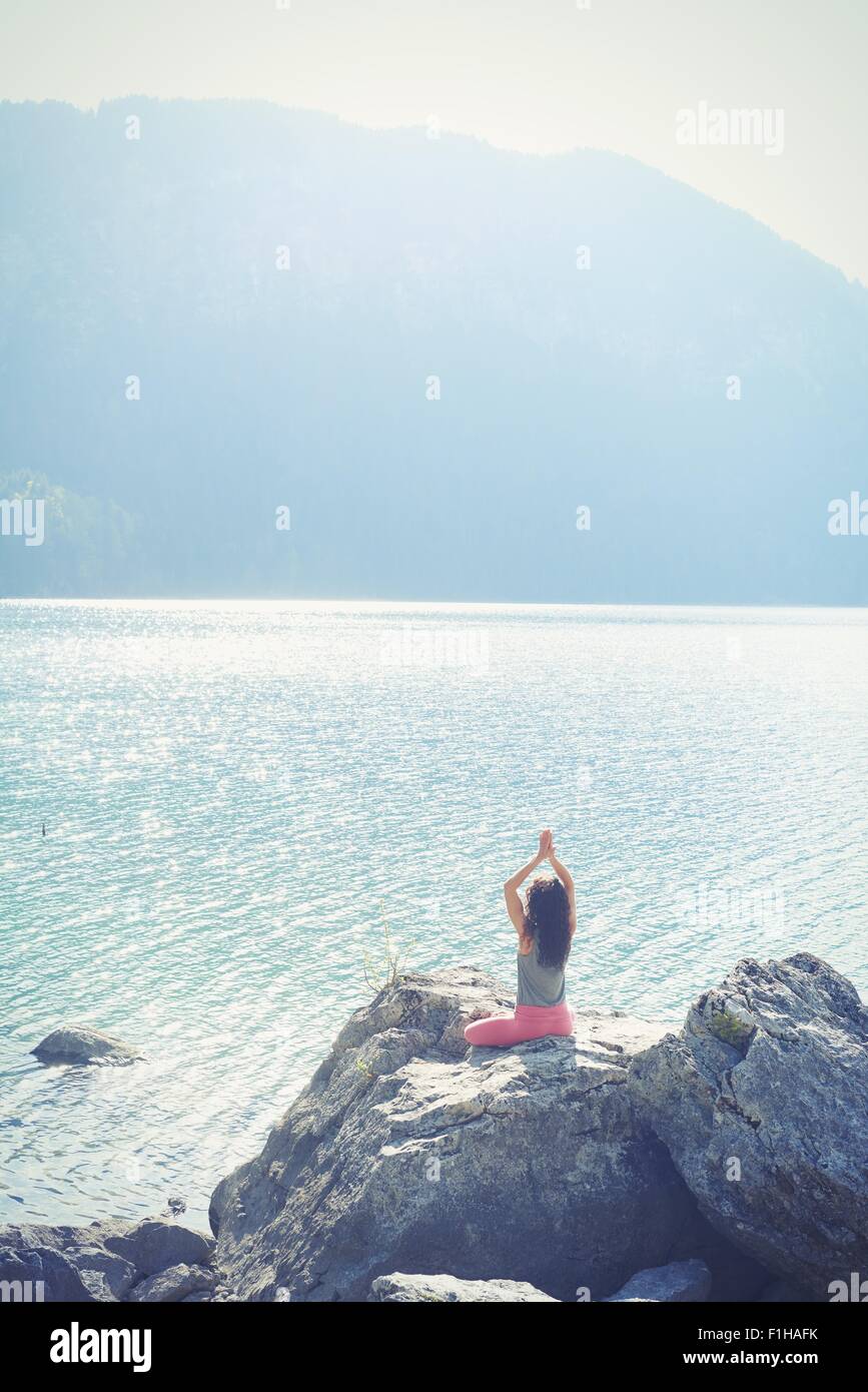 Mitte der erwachsenen Frau, sitzt auf Felsen am See in Yogaposition Rückansicht Stockfoto