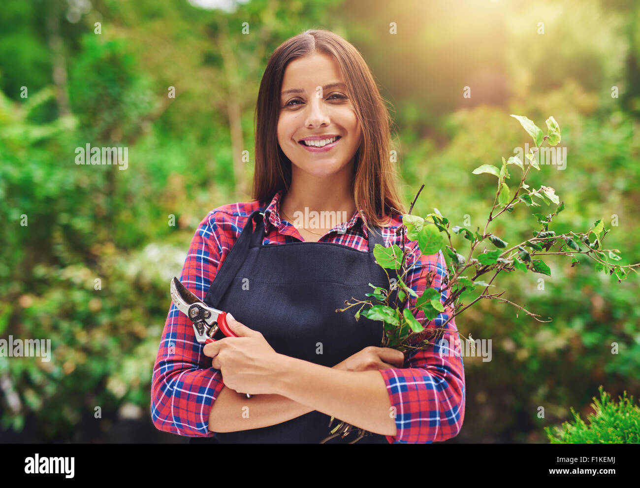 Lächelnde junge weibliche Gärtner Beschneiden der Pflanzen in einem üppigen Garten mit einem apir der Gartenschere in der Hand am Lächeln Stockfoto