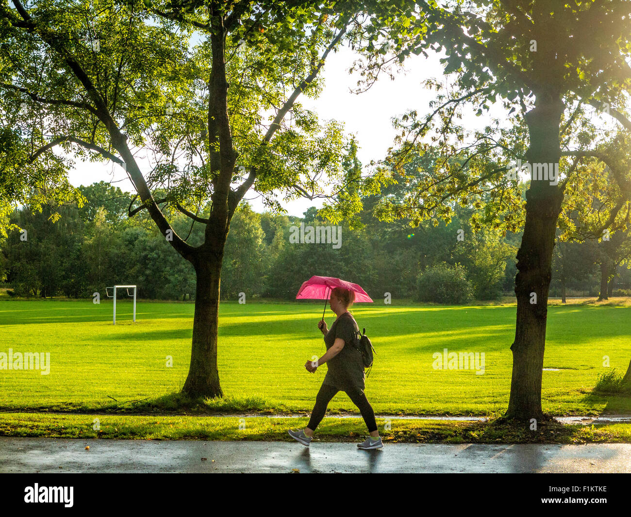 Eine Frau geht entlang der Straße mit einem hellen rosa Regenschirm, wie die Sonne nach einem Regenschauer kommt Stockfoto