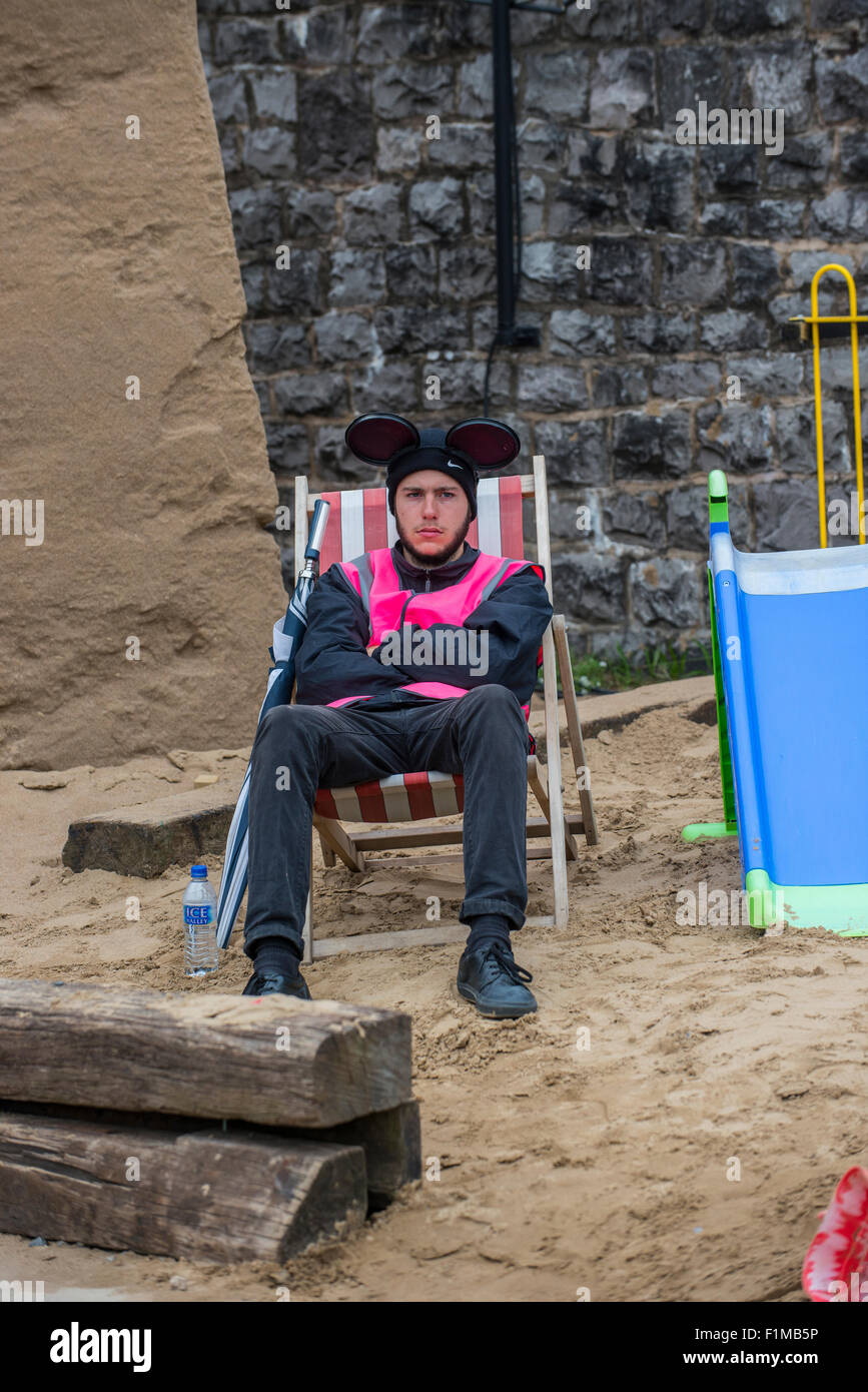 Angestellter bei Banksy Dismaland Verblüffung Park sitzt auf einer gefälschten Strand vor eine enorme Sandburg. Stockfoto