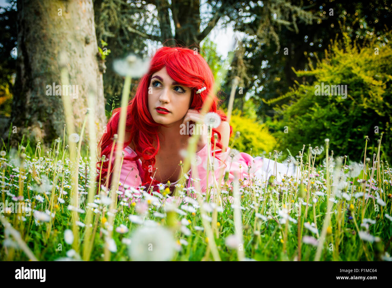 Junge Frau mit leuchtend roten Haaren liegen im Wald Blumenwiese mit Hand am Kinn Blick verträumt aus in den Weltraum Stockfoto