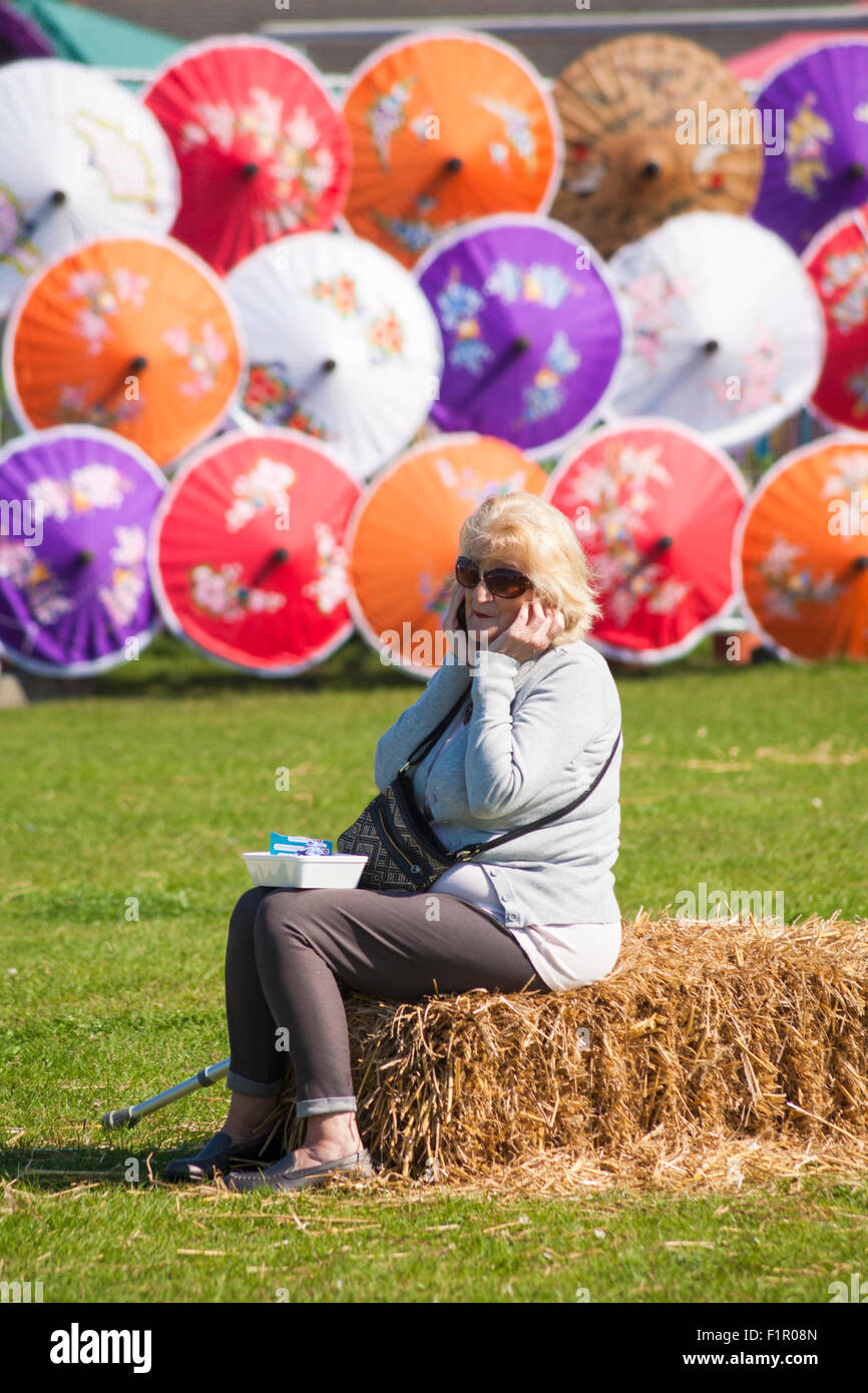 Poole, Dorset, UK. 6. September 2015. Poole Thai Festival etablierte sich als eines der beliebtesten Thai Festival des Landes. Frau am Telefon sitzen auf Heuballen mit bunten Sonnenschirmen hinter Credit: Carolyn Jenkins/Alamy Live News Stockfoto