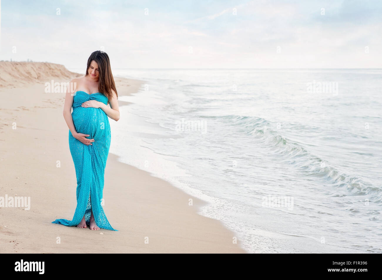 Schwangere Frau an einem Strand eine türkise blau Mutterschaft Kleid tragen. Sie lächelt und blickte auf ihren Bauch. Stockfoto