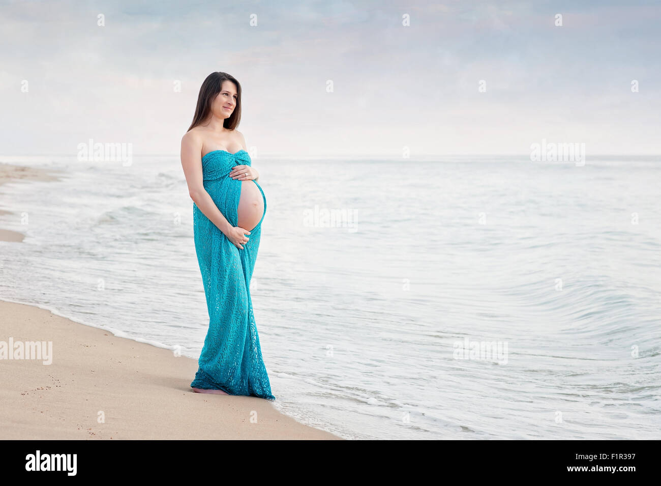Schwangere Frau trägt ein türkise blau Mutterschaft Kleid. Sie lächelt und mit Blick auf den Ozean. Stockfoto