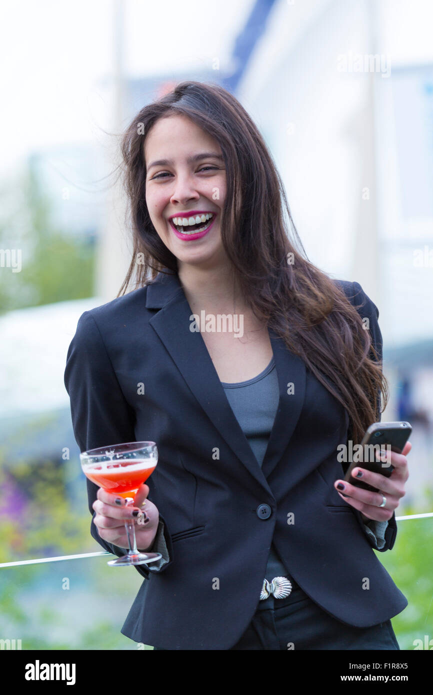 Junge lateinamerikanische/lateinamerikanische Brünette, lächelnd, mit Handy und Getränk Stockfoto