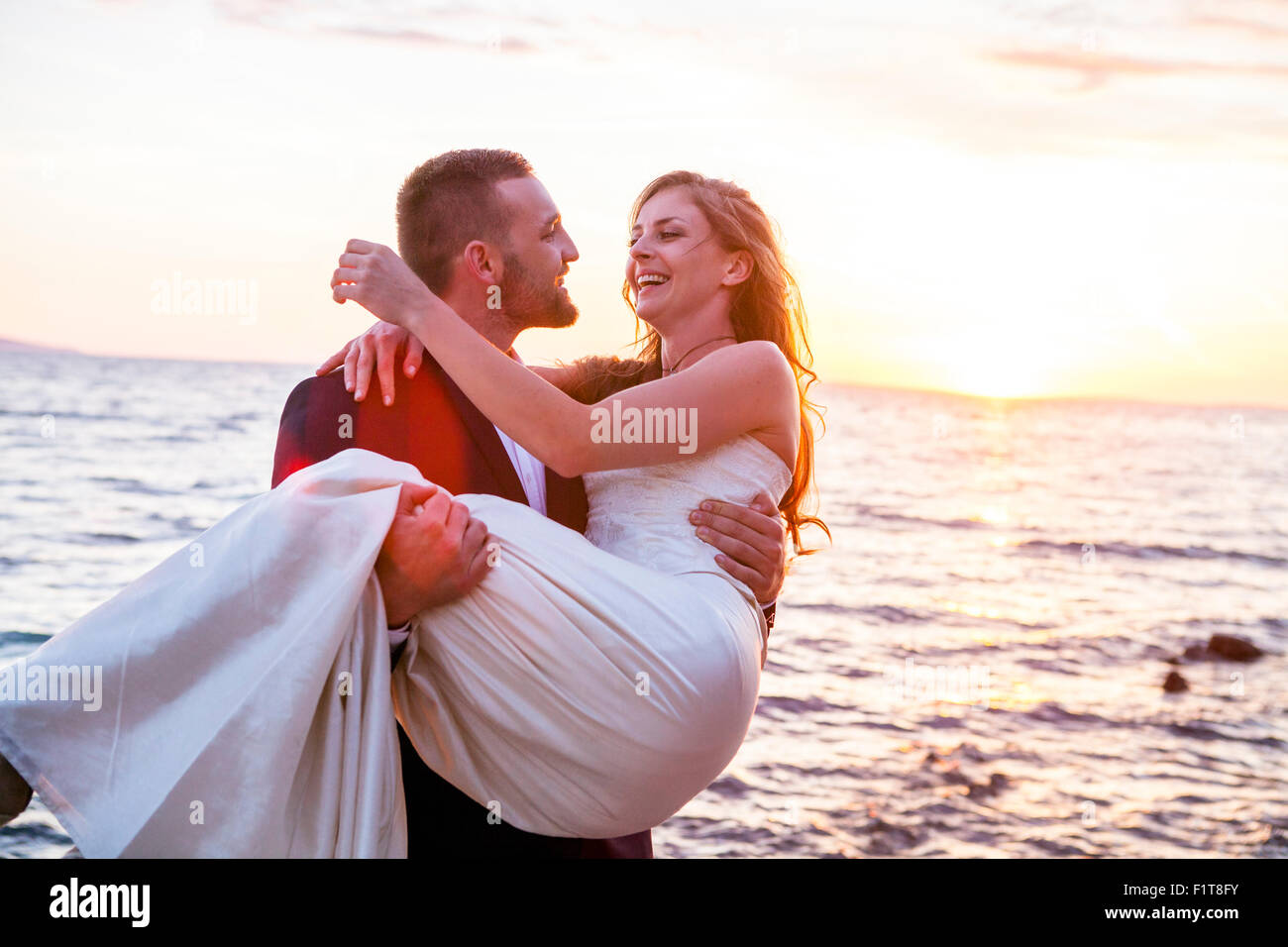 Bräutigam trägt Braut am Kiesstrand bei Sonnenuntergang Stockfoto