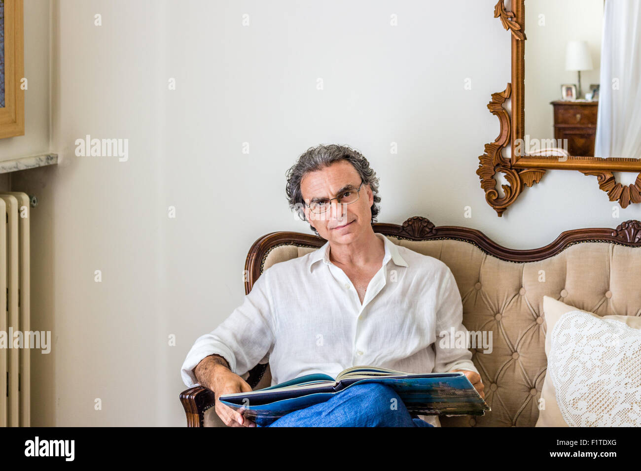 faszinierende vierziger ist Mann raffiniert und doch sportlich gekleidet eine Brille und einen Foto-Bildband über ein gepolstertes Sofa in eine helle und luxuriöse Wohnzimmer beobachten Stockfoto