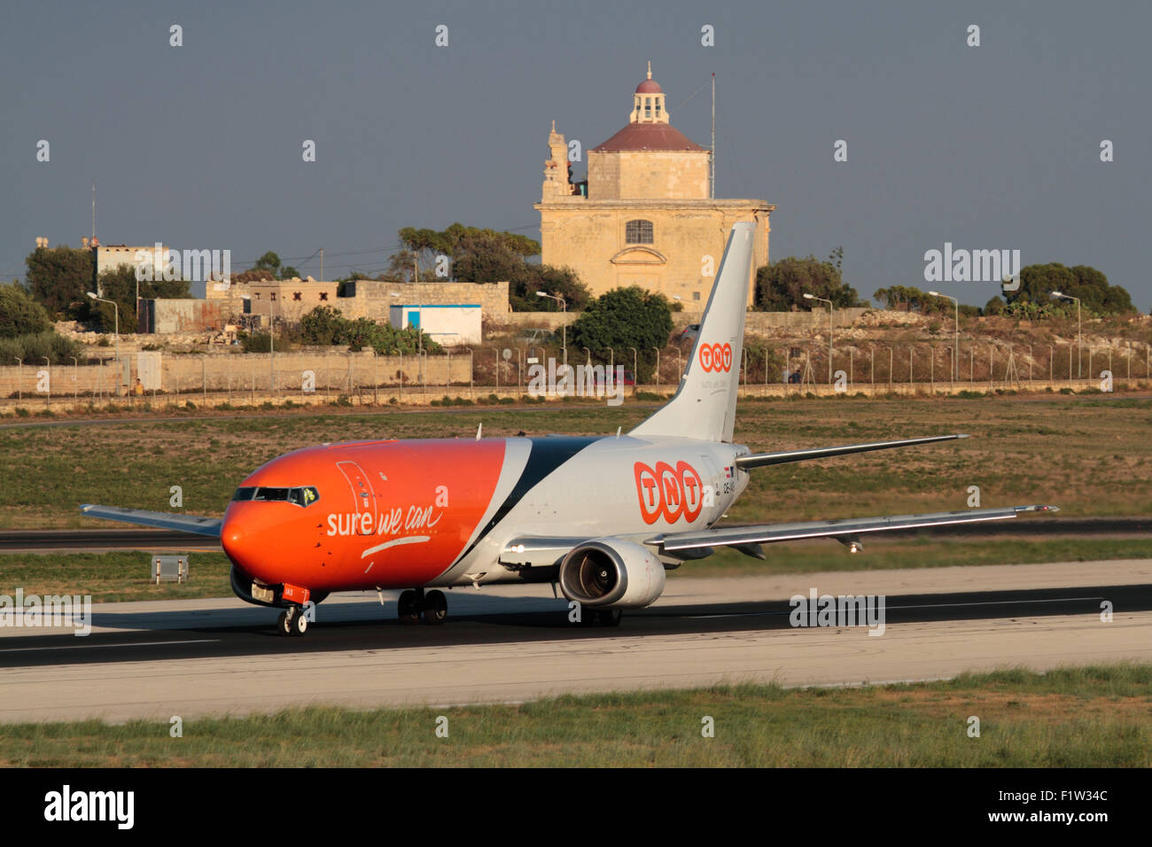 TNT Airways Boeing 737-400 Frachtflugzeug ausziehen aus Malta mit Ta' Loretu Kapelle, Gudja, im Hintergrund Stockfoto