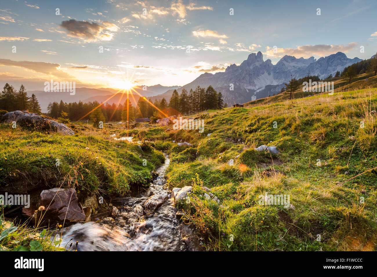 Die Sonne geht hinter den Bergen und wirft das letzte Licht auf eine hohe Weide mit einem kleinen Gebirgsbach. Stockfoto