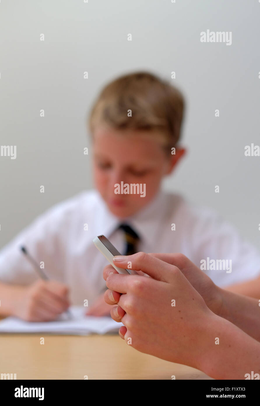 Ein Schüler mit einem Mobiltelefon (SMS) in der Klasse wie ein weiterer Schüler im Hintergrund arbeitet Stockfoto