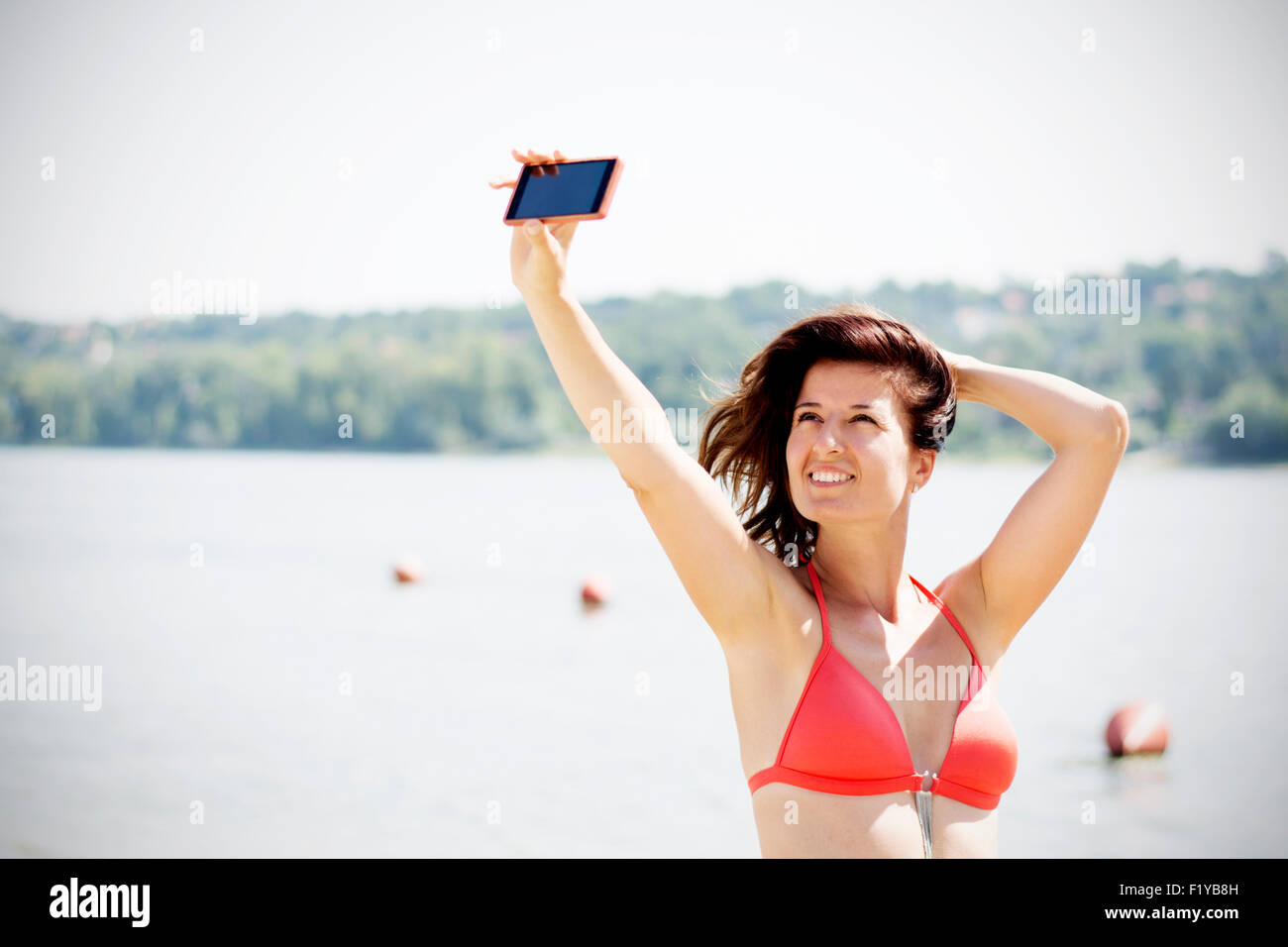 Attraktive junge Brünette Frau Selbstbildnis mit ihrem Handy am Strand nehmen. Sie trägt rote Bikini und lächelnd. Stockfoto