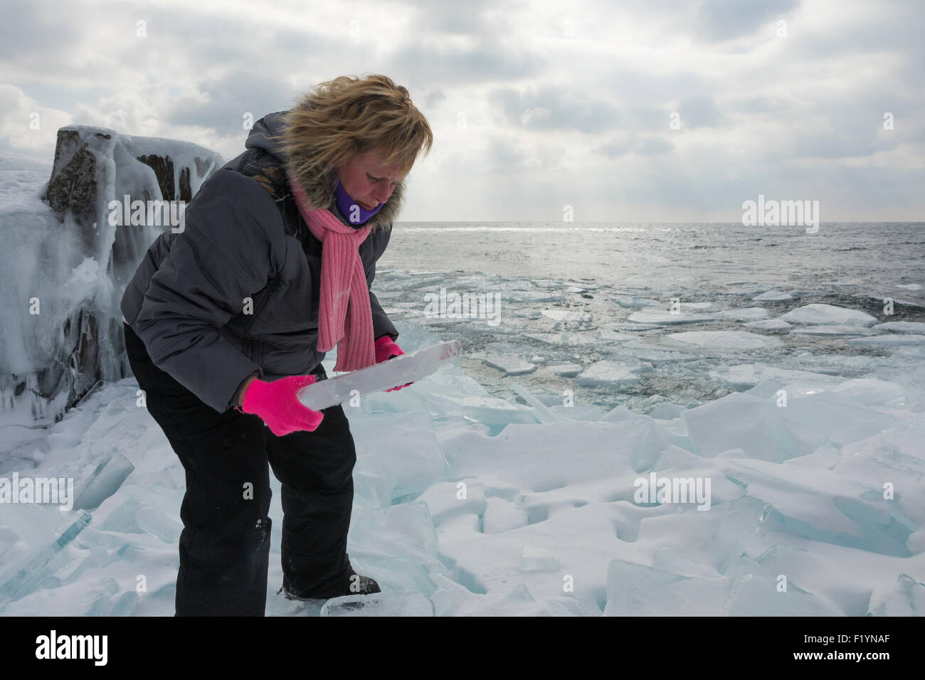 Blonde Frau kaukasischen in ihren 40ern trägt einen Wintermantel und rosa Schal nimmt ein großes Stück der Platte Eis am Lake Superior Stockfoto