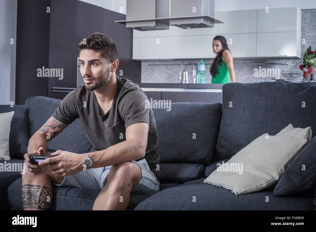 Junger Mann auf Sofa mit Gaming Control beobachtet von böse Freundin Stockfoto