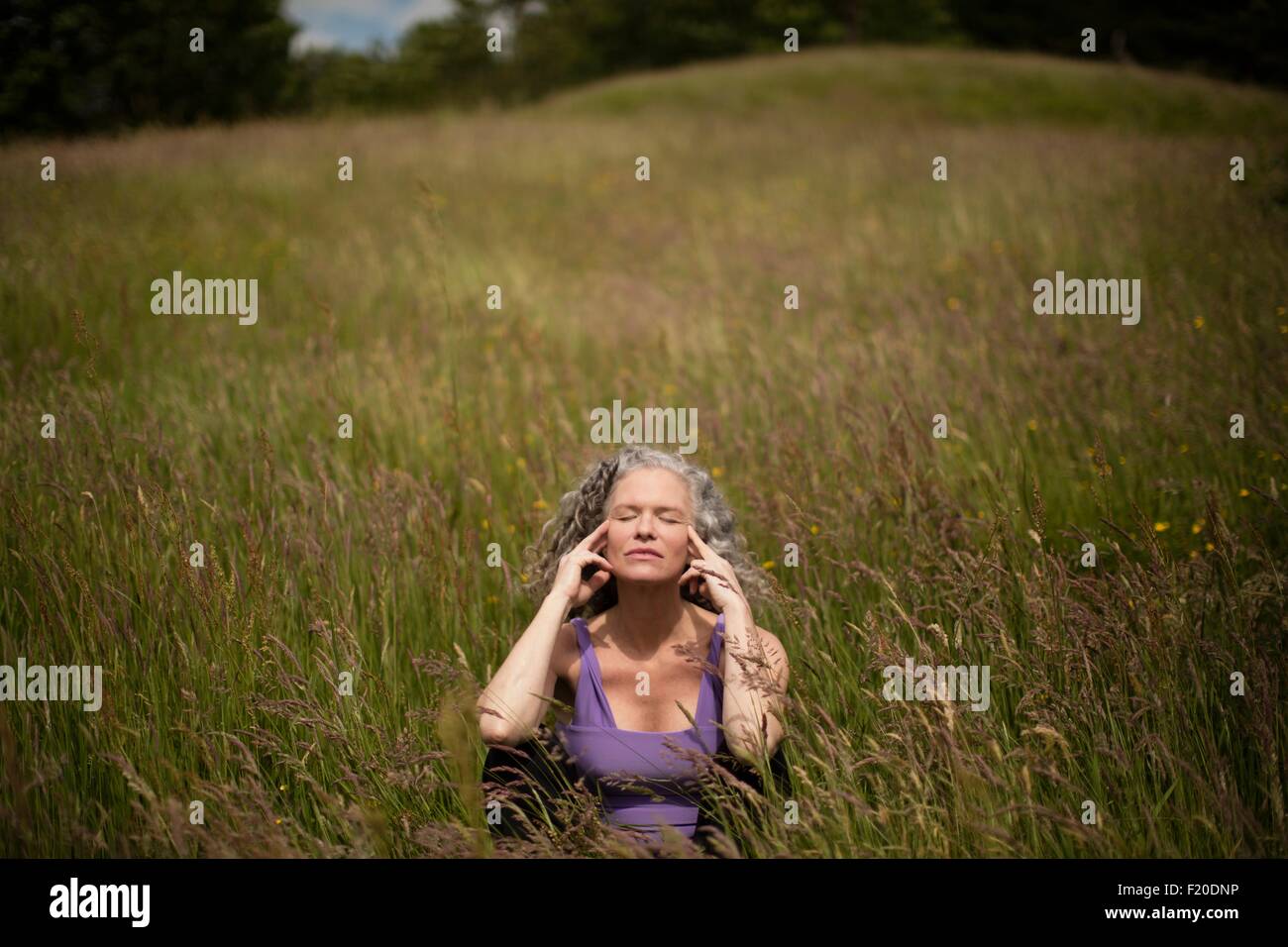 Reife Frau mit Fingern auf ihre Tempel sitzen in langen Rasen zu meditieren Stockfoto