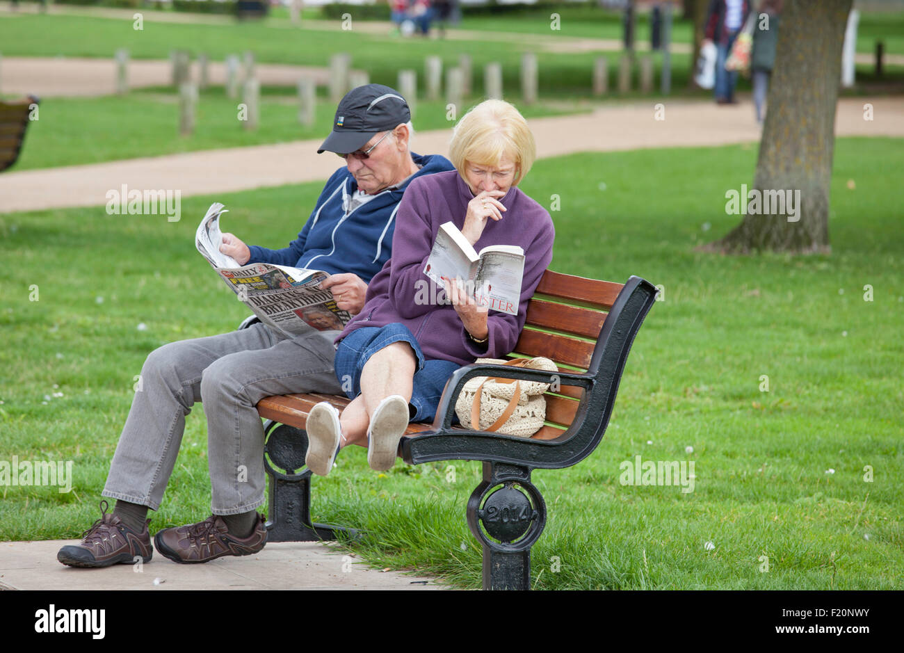 Applying paar, entspannend, die Dame mit einem Taschenbuch und der Mann mit einer Zeitung auf einem Park-Sitz, England, UK Stockfoto