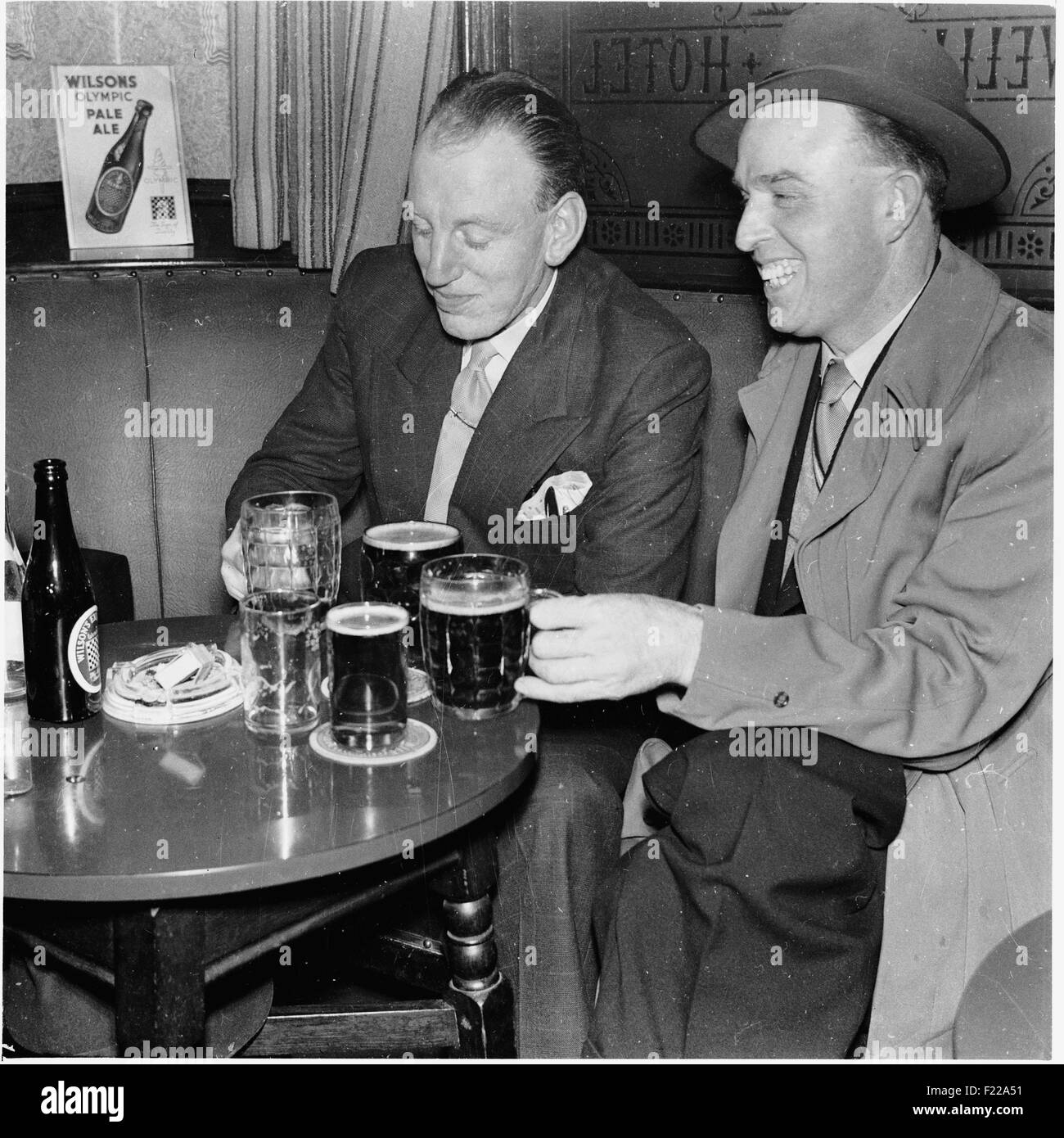 Geschichtsbild aus den 1950er Jahren, zwei Männer geeignet, eins mit einem Regenmantel und trägt einen Hut, sitzen zusammen in einem Pub genießen Sie mehrere Gläser und Flaschen von Wilsons pale Ale, Manchester, UK. Stockfoto